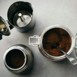 A flat lay image of a moka pot, ground coffee, and jar on a textured gray background.